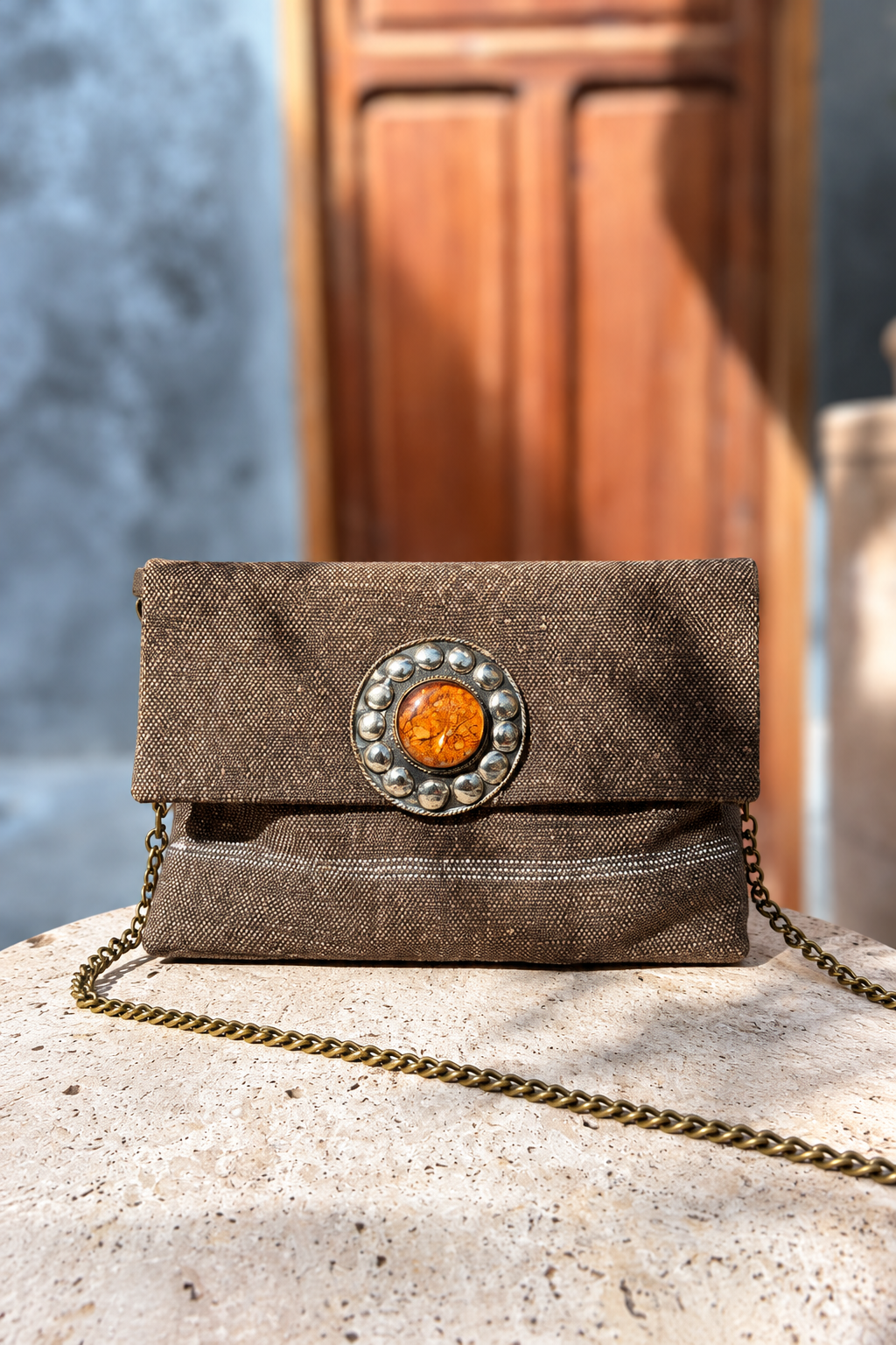 Legizta Earthy tribe brown carpet bag with orange centrepiece and chain strap on a beige stone pedestal in front of a wooden doorway
