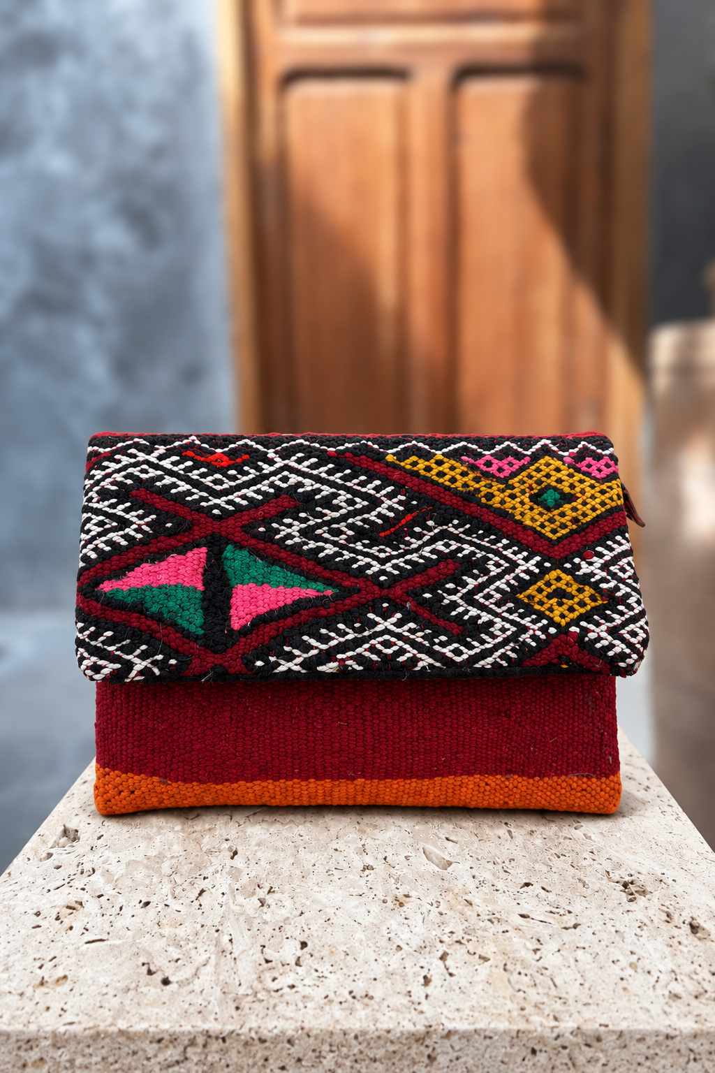 Legzita Nemo carpet bag with colourful geometric embroidered flap and burgundy body on a beige stone pedestal in front of a wooden doorway